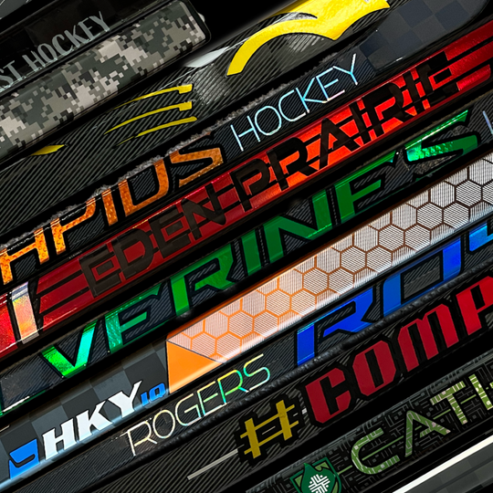 A close-up view of stacked hockey sticks, each featuring colorful branding, team logos, and customizable hockey stick graphics from various manufacturers.